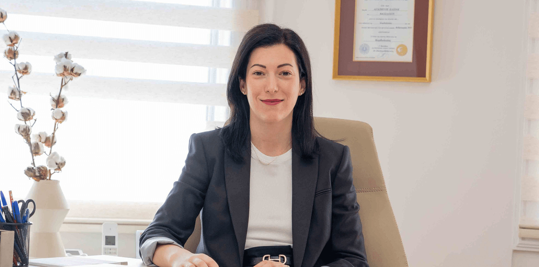 Η γιατρός καθισμένη στο γραφείο της, φοράει κοστούμι και κοιτάζει χαμογελαστή την κάμερα σε φωτεινό επαγγελματικό περιβάλλον