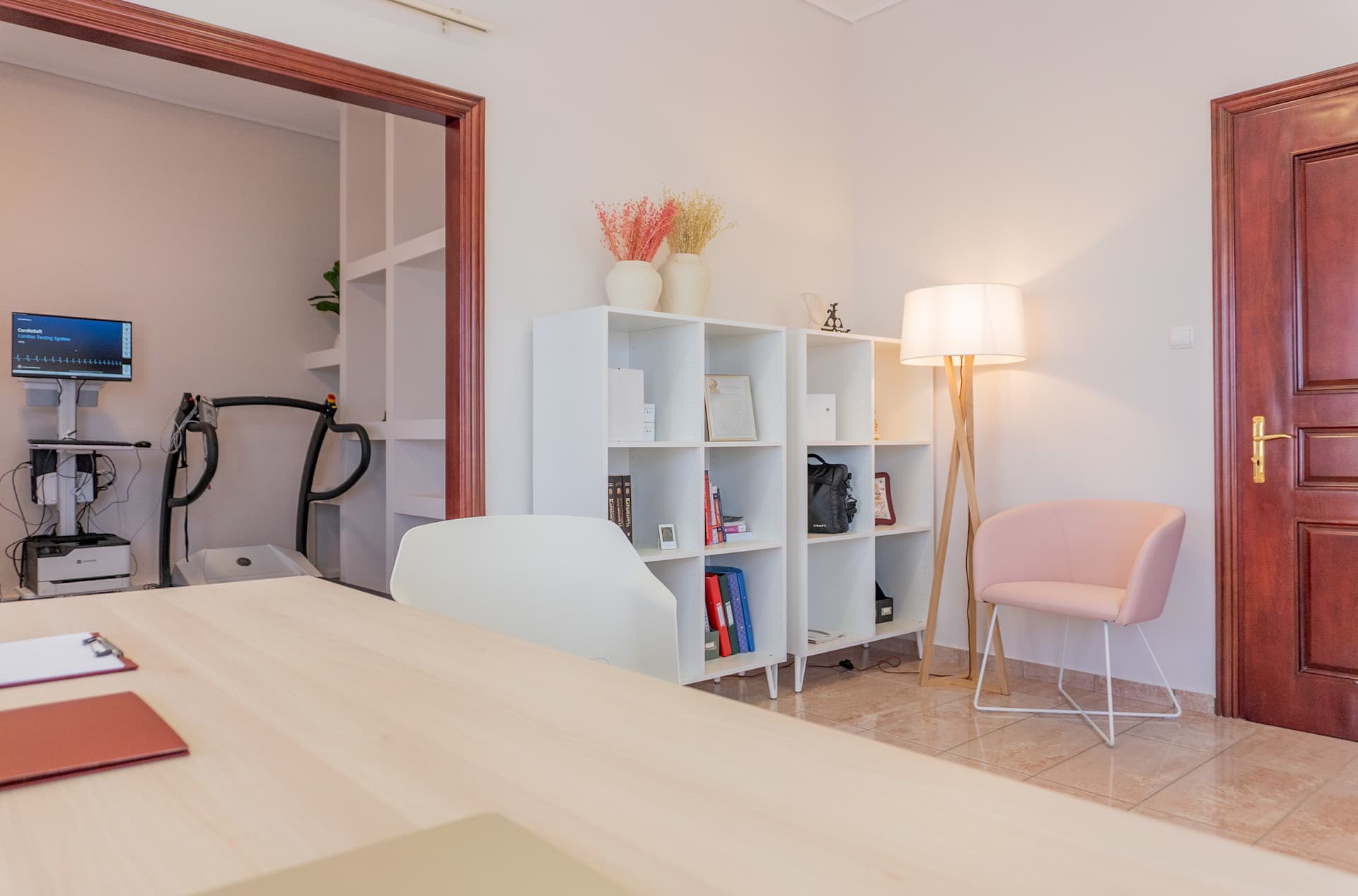 Γραφείο της ιατρού με βιβλιοθήκη, διακόσμηση και καρέκλα αναμονής. Ο εξοπλισμός του εξεταστηρίου είναι ορατός στο βάθος.